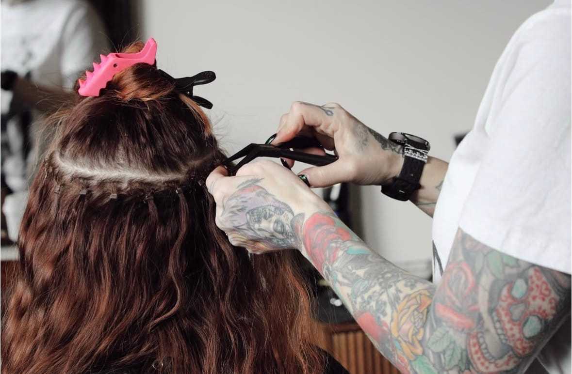 Tattooed stylist attaching hair extensions to a client's red hair using clips and a comb.