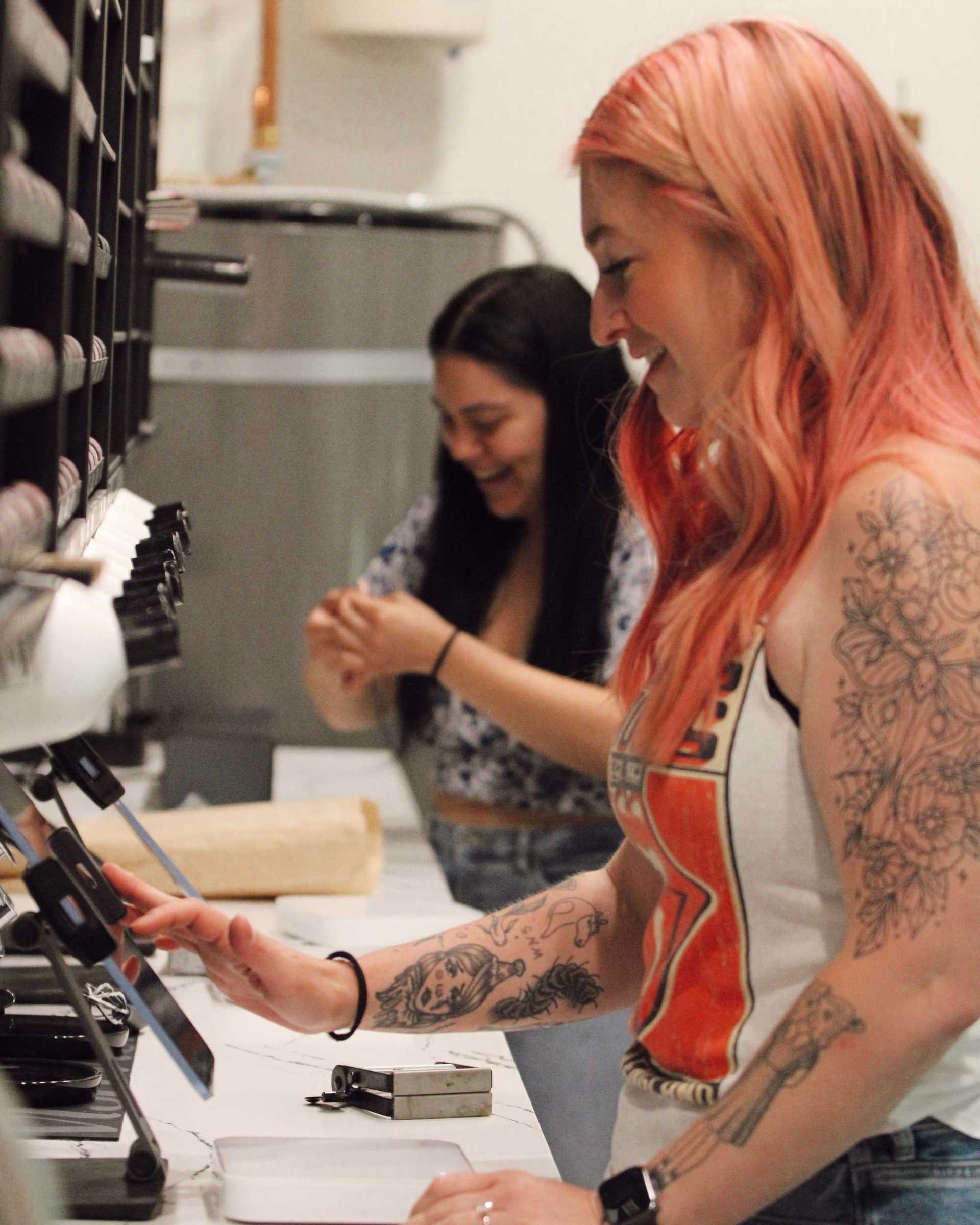 Two women with tattoos working in a print shop, smiling and engaged in their tasks.