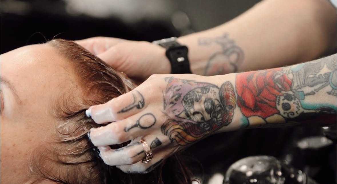 Person with tattoos washing another's hair at a salon.