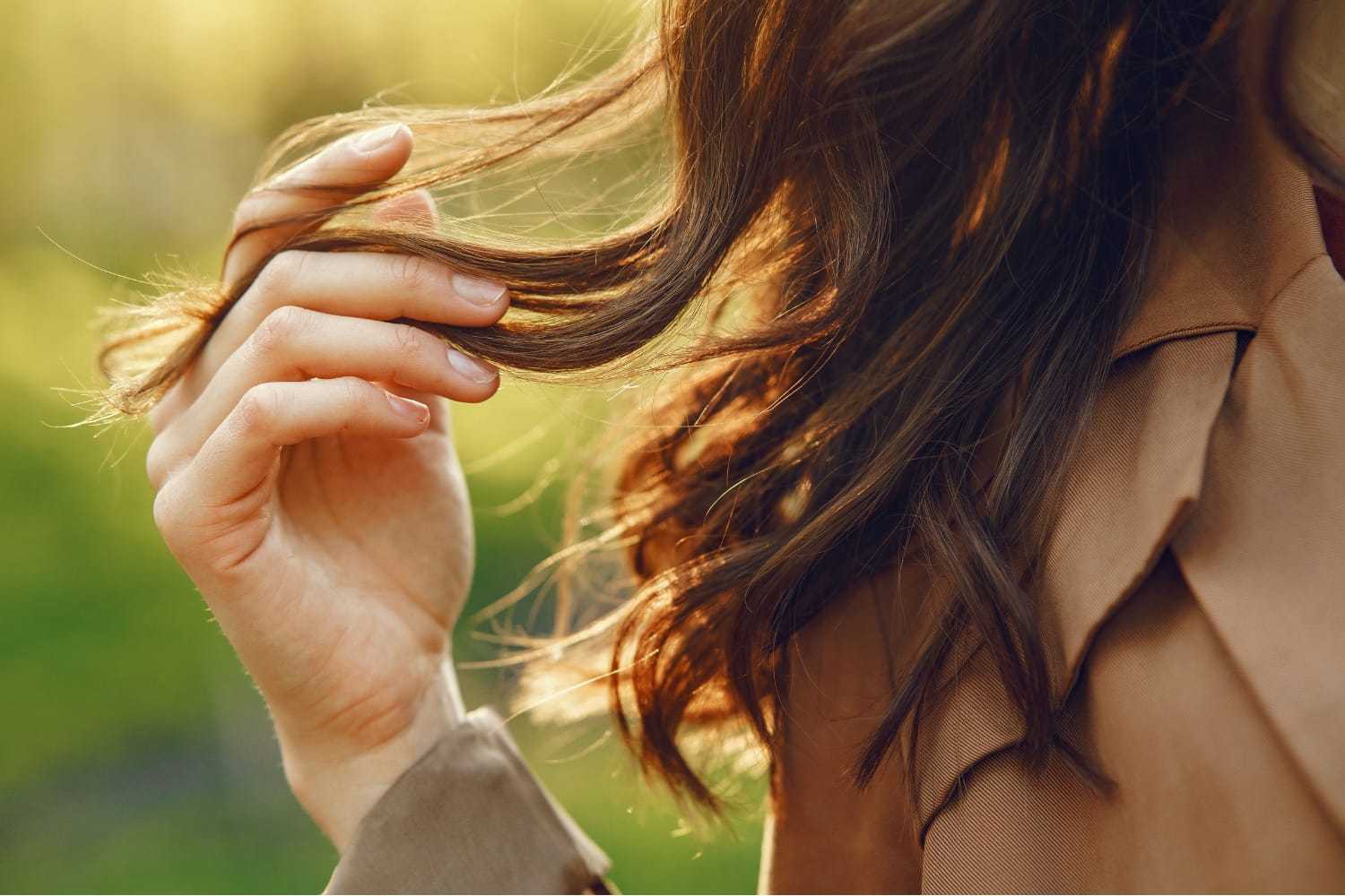 Close-up of a woman's hand holding flowing brown hair, with blurred greenery in the background.