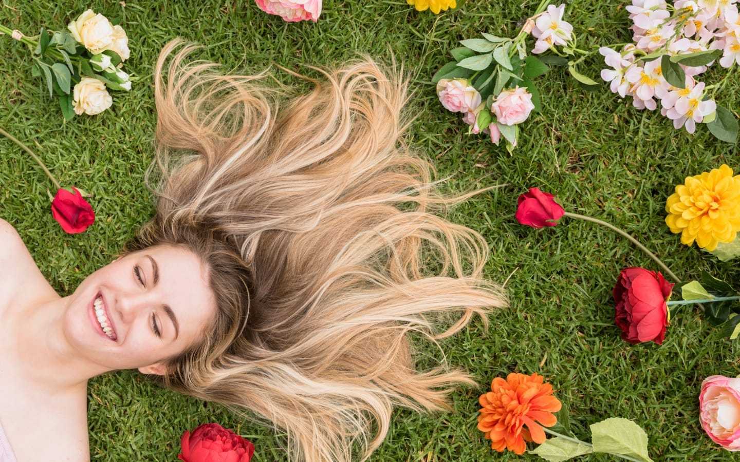 Smiling woman lying on grass with scattered colorful flowers around her.