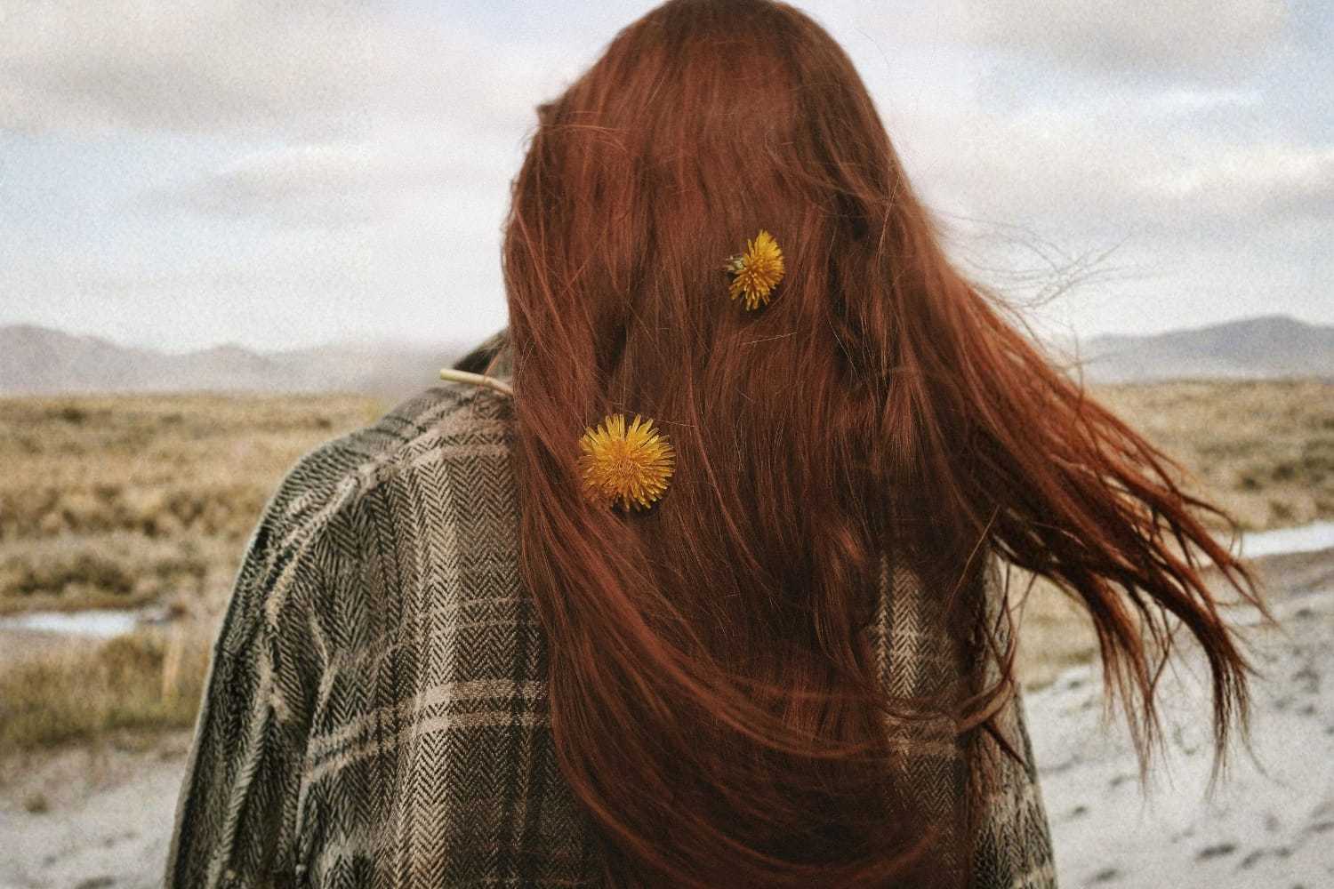A woman with long red hair adorned with yellow flowers faces a vast, windy landscape.