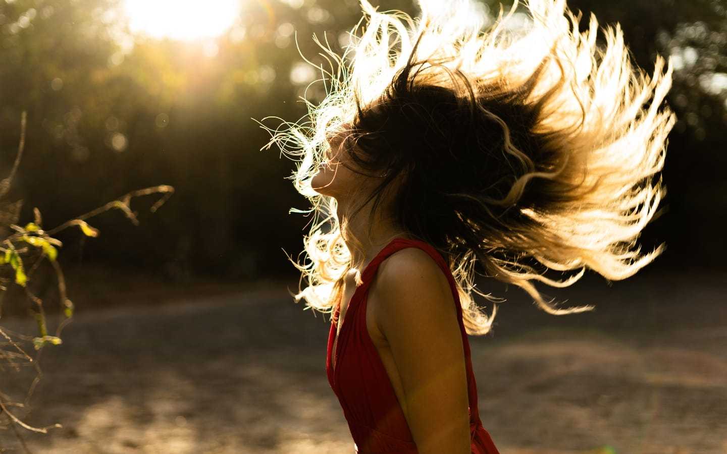 Woman in red dress with flowing hair, backlit by sunlight in an outdoor setting.