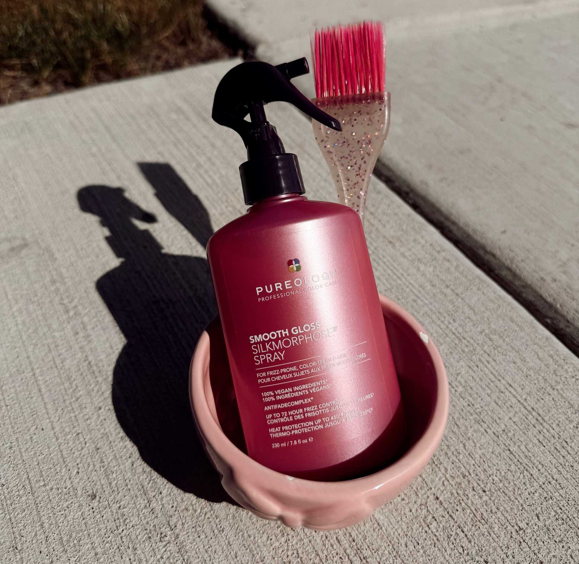 Bottle of silk morph gloss spray in a pink bowl, next to a pink-tipped brush on concrete.